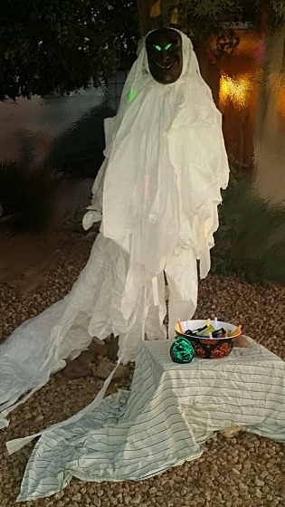 A white figure with a dark wood mask and glowing green eyes, stands guard over a small table draped with a sheet and bearing a black wire lantern filled with green LED lights, and a bowl of candy and lit glowsticks.