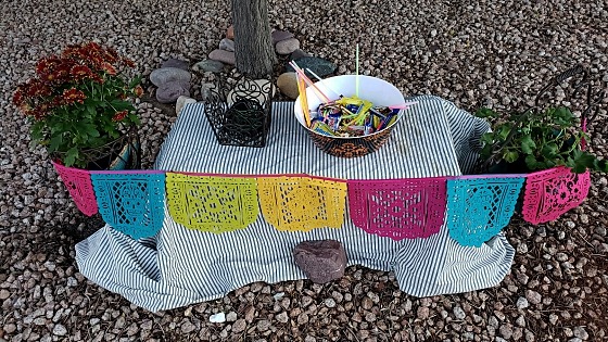 A low table with a light-colored striped sheet, a draped pieced felt Dia de los Muertos banner in bright colors, a scrollwork wire lantern, and a plastic bowl filled with candy and lit glowsticks.