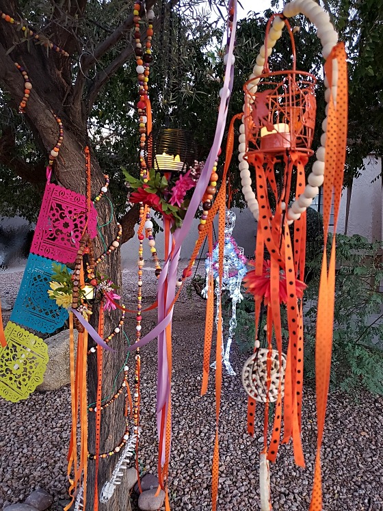 Daytime photo of my 2023 Halloween display, with bright colored whood bead garlands, pieced felt banners, fluttering ribbons, and wire lanterns hung with flowers and beads.