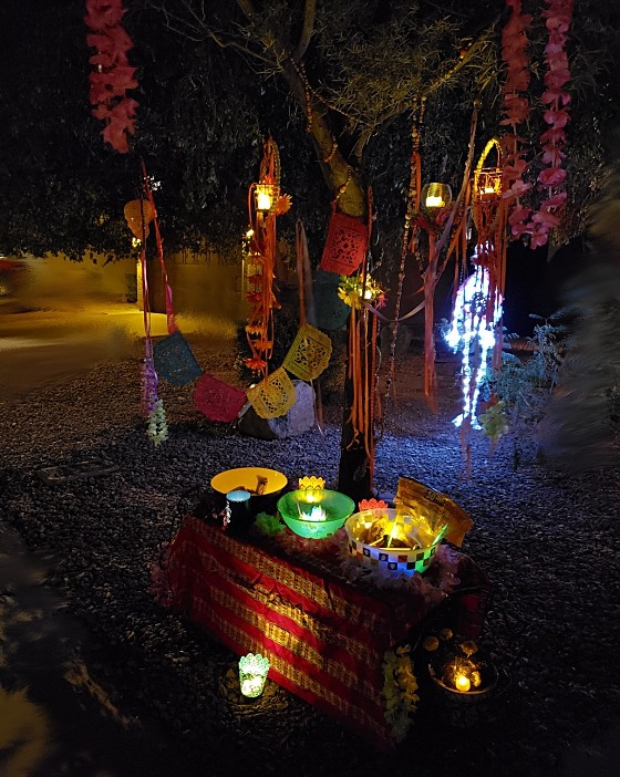 A shot of the ofrenda table at night, with bowls of candy and glowsticks lit by lanterns draped in silk flower garlands