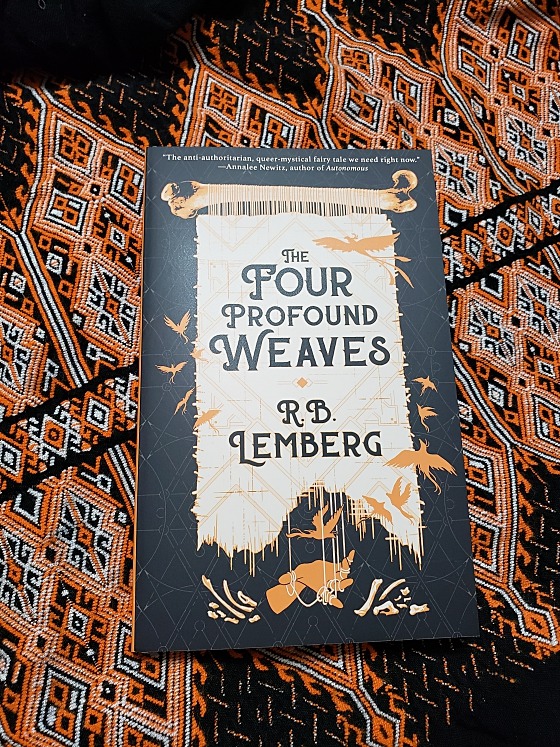 The cover to R. B. Lemberg's 2020 book THE FOUR PROFOUND WEAVES, on a backdrop of orange and white geometric weaves on black cotton fabric.